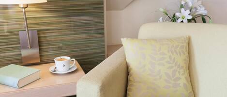 A hotel room with a sofa, a bedside table with a lamp, a cup of coffee, and a flower arrangement.