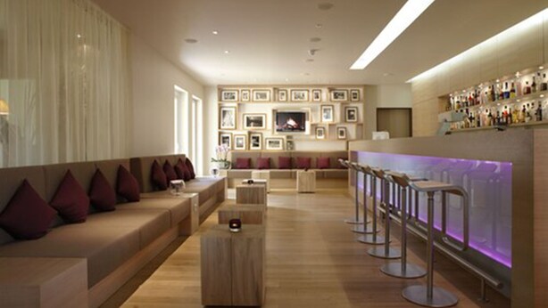 A modern bar area with a long counter and multiple stools. There is a seating area with a sofa and cushions. The room has a neutral color palette and features framed pictures on the walls.