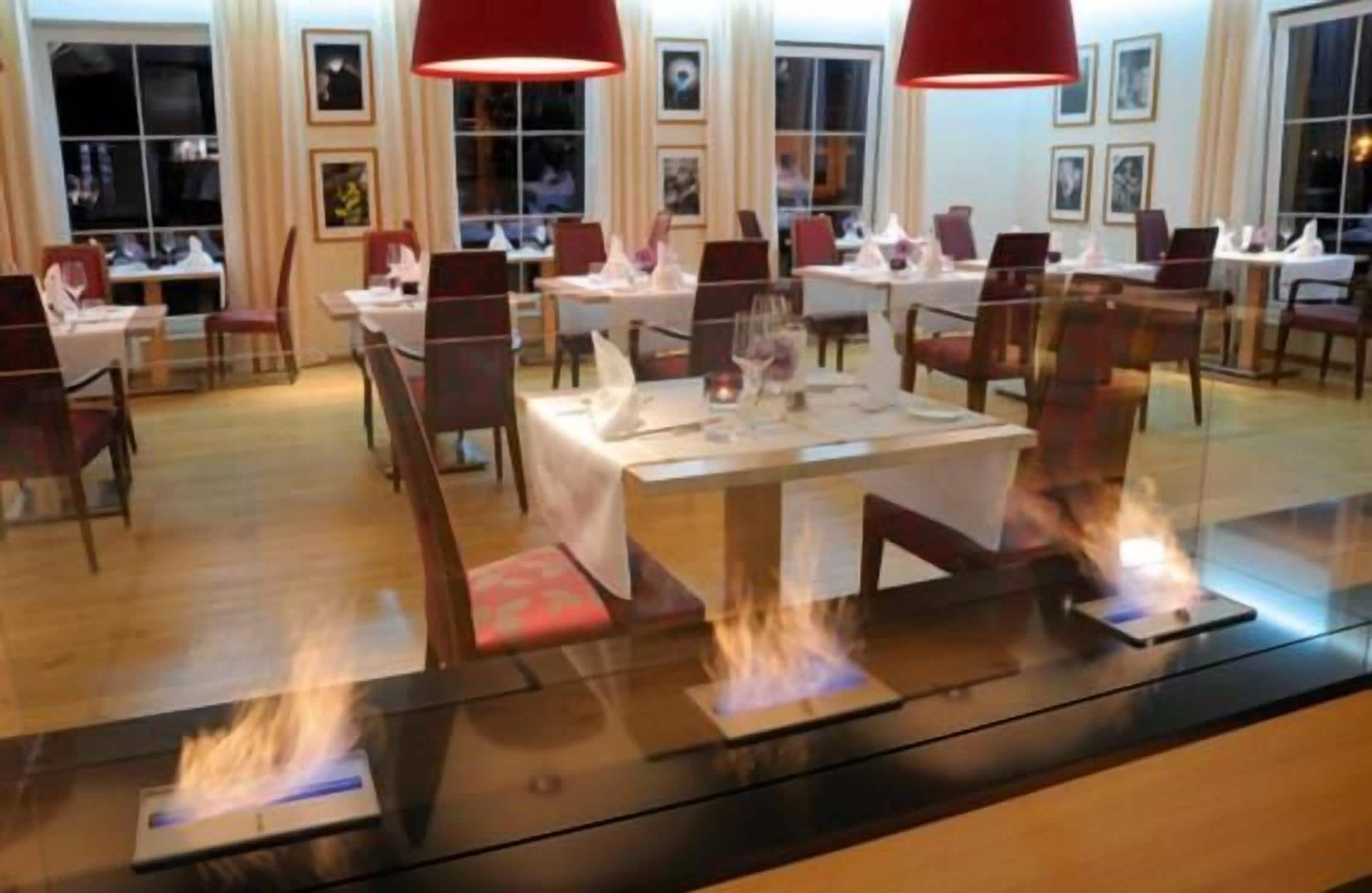 A restaurant with tables set for dining, overhead red lighting, and a central fire feature.