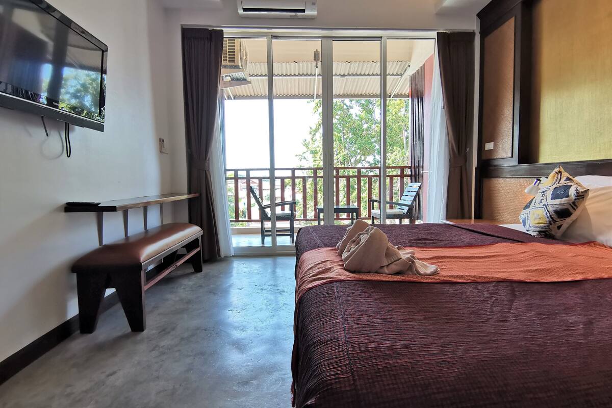 A hotel room with a bed, a bench, a TV, and a balcony with chairs.