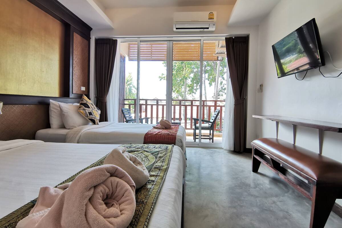 A hotel room with two beds, a TV, a balcony with a chair, and an air conditioning unit.