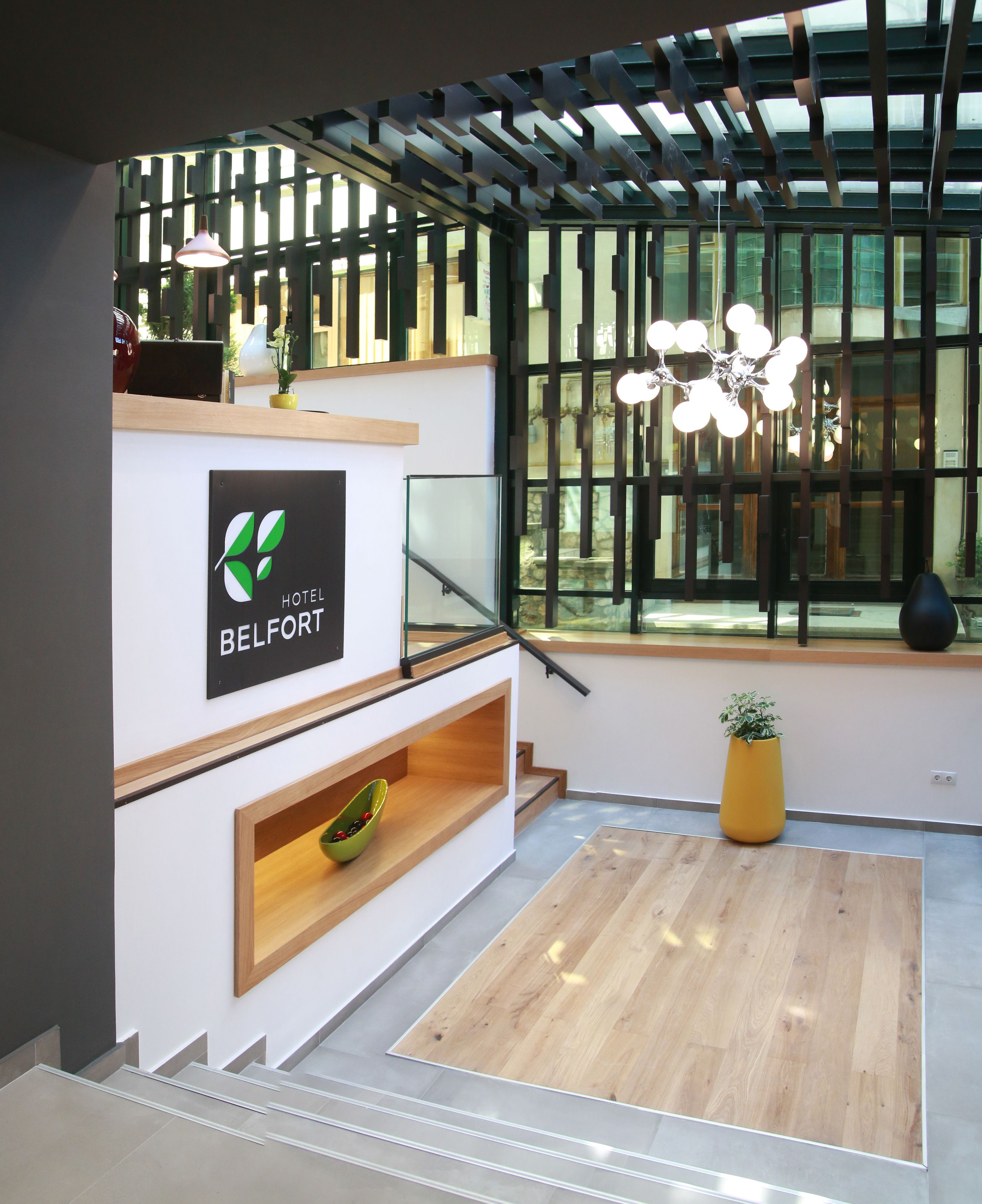 A modern hotel lobby with a reception desk, a staircase, and a large window with natural light.