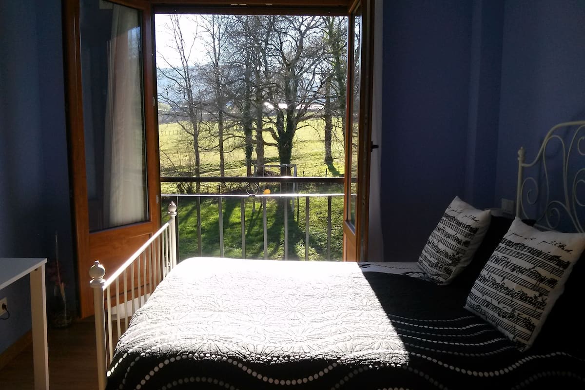 Een slaapkamer met een bed, kussens, een raam met uitzicht op bomen en een balkonhek.