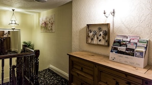 A hallway with a wooden staircase, a mirror, a framed floral artwork, and a display of travel brochures.