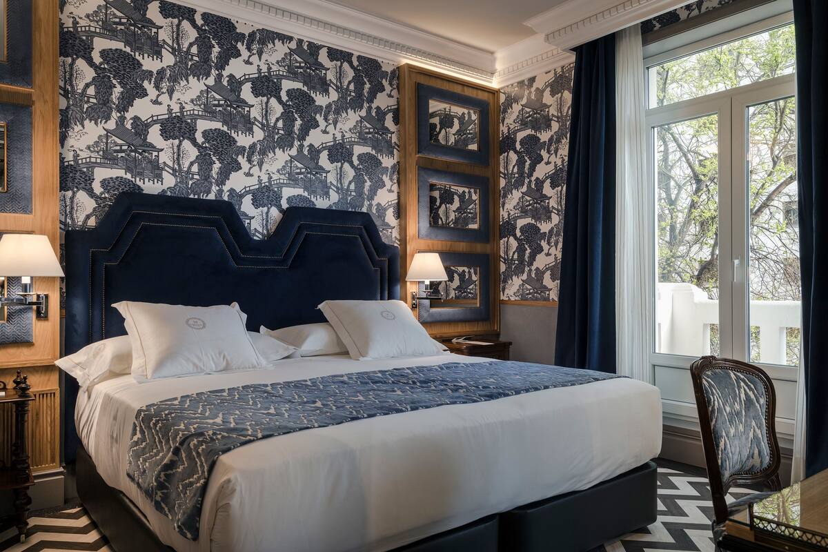 Een opgemaakt bed met een donker hoofdbord, witte beddengoed en een gestreept plaid. Er is een raam met gordijnen, een stoel en een muur met een gedetailleerde muurschildering.