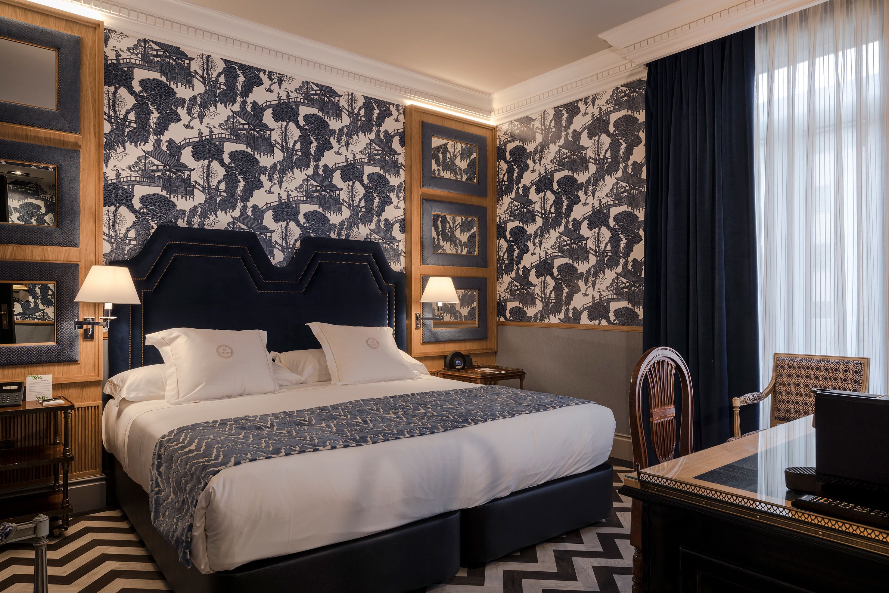 Quarto de hotel com uma cama grande, uma escrivaninha e uma cadeira. Existem duas luminárias nas mesas de cabeceira, um espelho e uma janela com cortinas.