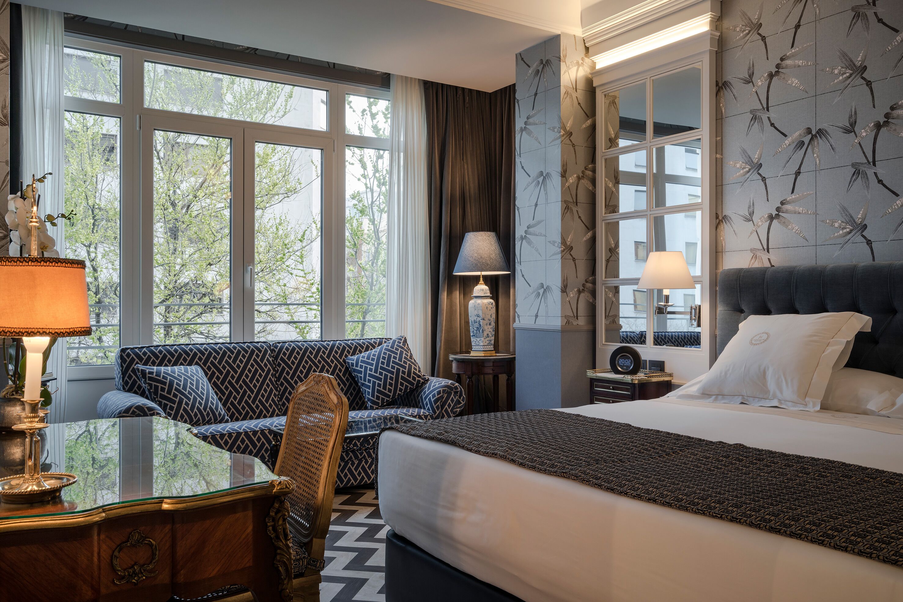 A hotel room with a large bed, a sofa, a desk with a lamp, and a view of trees through the windows.
