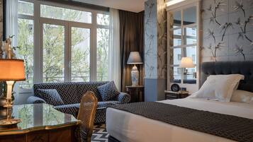A hotel room with a large bed, a sofa, a desk with a lamp, and a view of trees through the windows.
