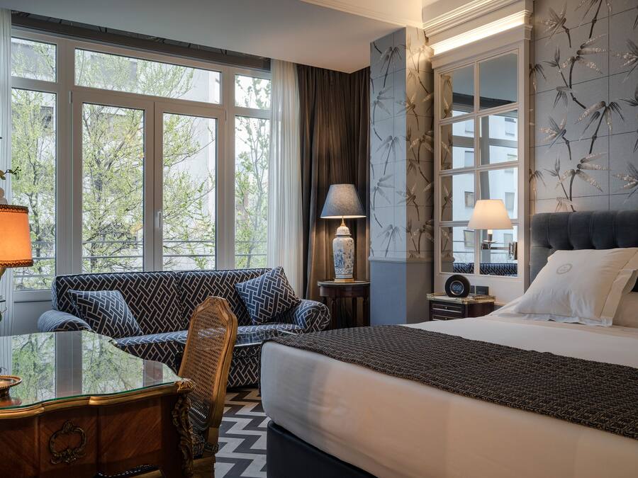 A hotel room with a large bed, a sofa, a desk with a lamp, and a view of trees through the windows.