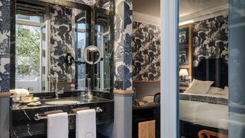 Un baño con lavamanos de mármol negro, un espejo grande y una ventana. En la habitación contigua hay una cama con ropa de cama blanca.