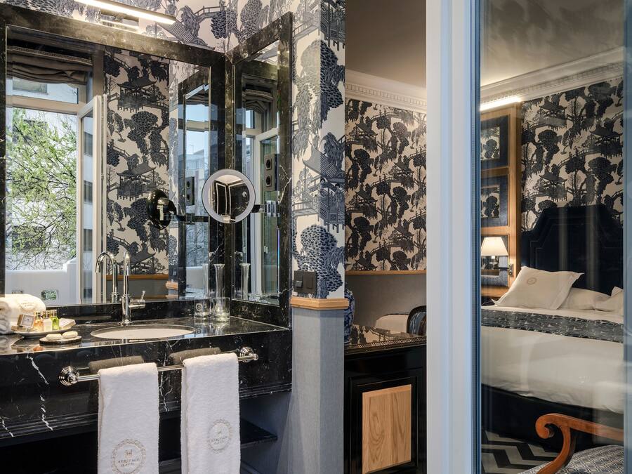 Un baño con lavamanos de mármol negro, un espejo grande y una ventana. En la habitación contigua hay una cama con ropa de cama blanca.