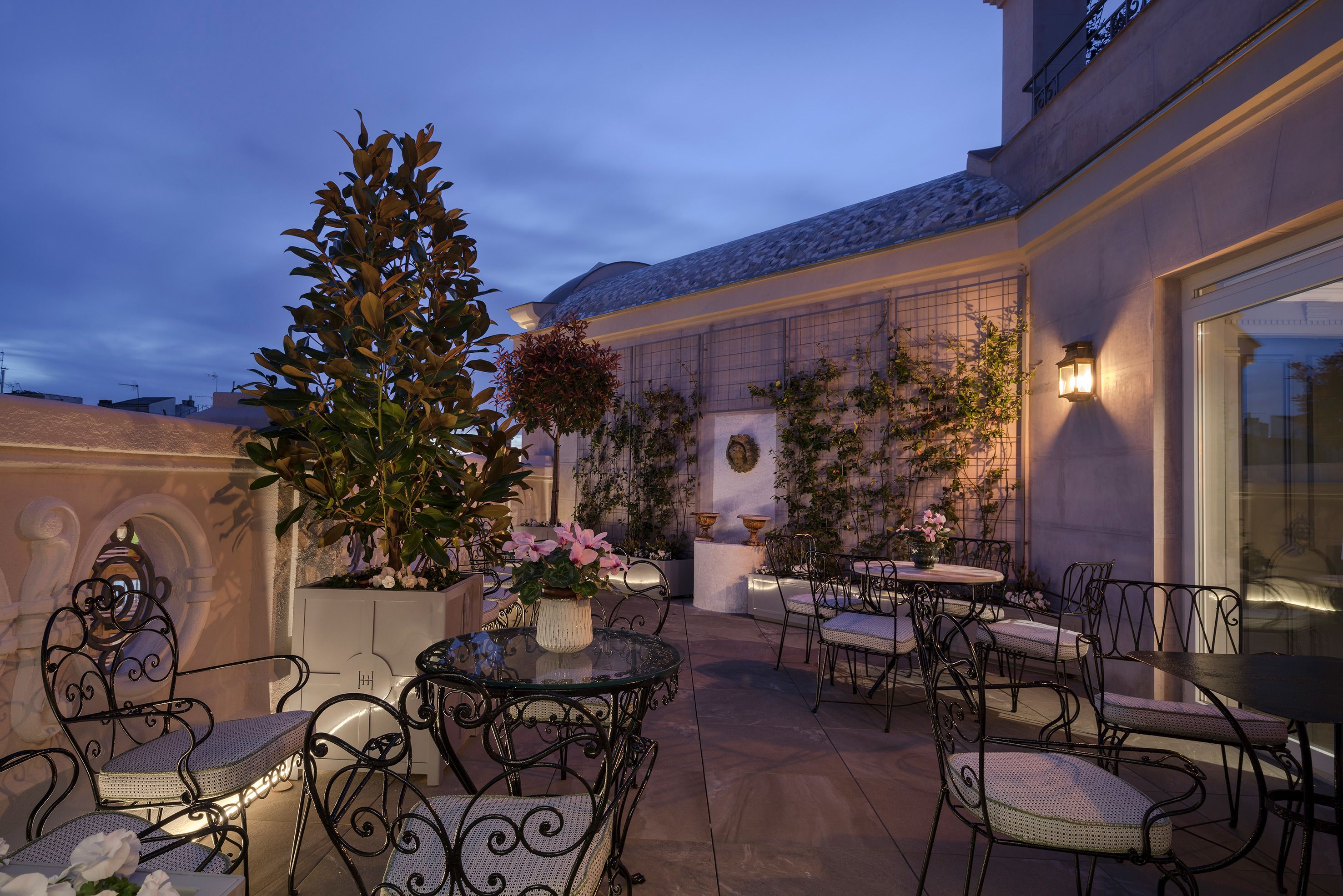 An outdoor patio area with round tables and chairs, surrounded by potted plants and decorative lighting.