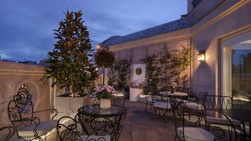 An outdoor patio area with round tables and chairs, surrounded by potted plants and decorative lighting.