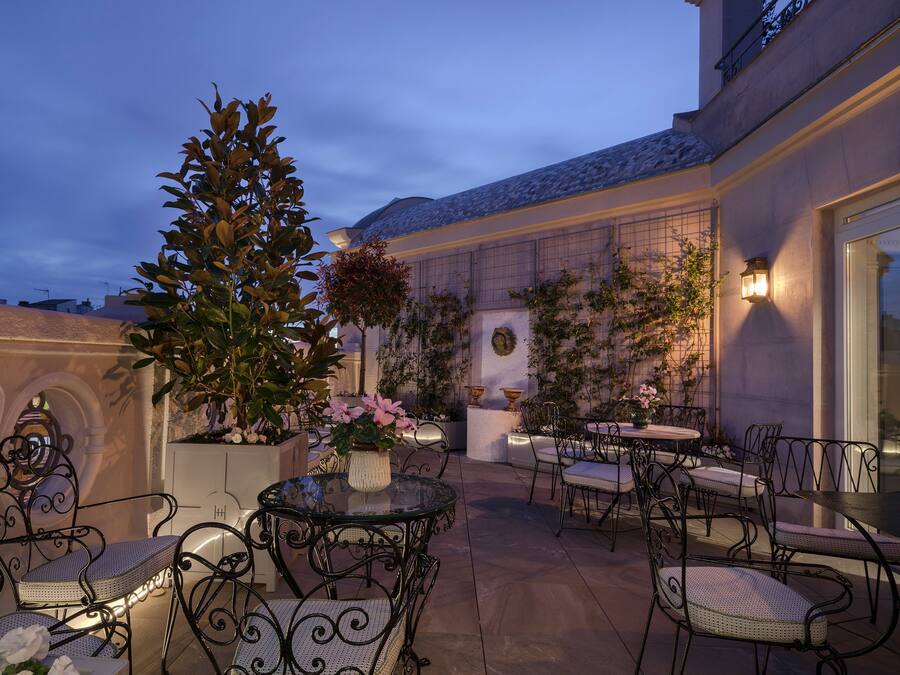 An outdoor patio area with round tables and chairs, surrounded by potted plants and decorative lighting.