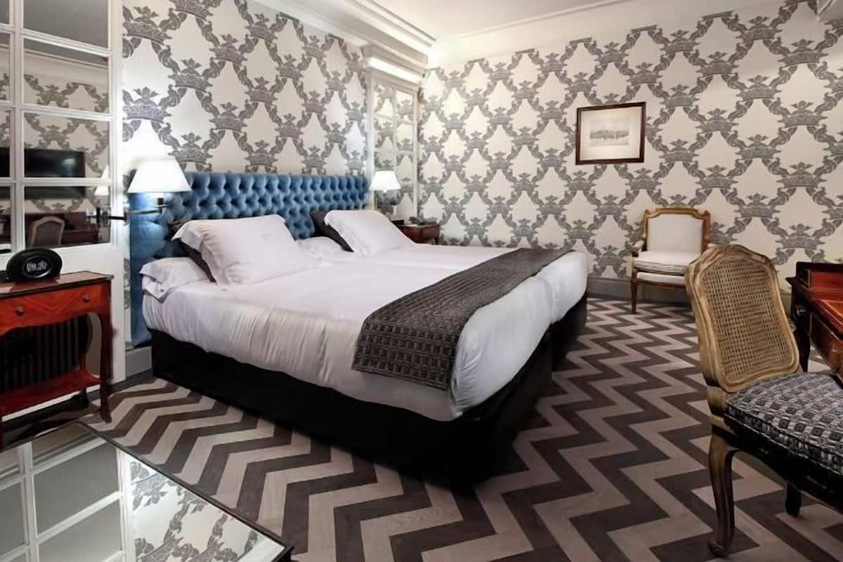 Een slaapkamer met een gestoffeerd hoofdeinde, een bed met witte lakens, een patroonrijk vloerkleed, een stoel en een dressoir.