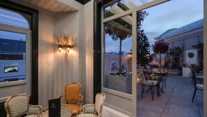 Un espace de détente intérieur, bien éclairé, avec vue sur la terrasse extérieure. On y trouve trois chaises et une table sur laquelle se trouve une boîte noire.
