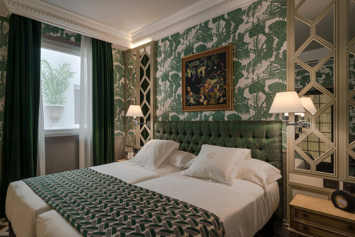 Een hotelkamer met een groot bed, twee bedlampen, een muur met een groen patroon en een raam met gordijnen.