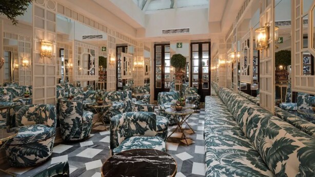 Un hall d’entrée spacieux doté d’un sol à carreaux, de coins salon avec des meubles recouverts d’un tissu à motifs de feuilles tropicales, et d’un grand miroir qui reflète l’espace.