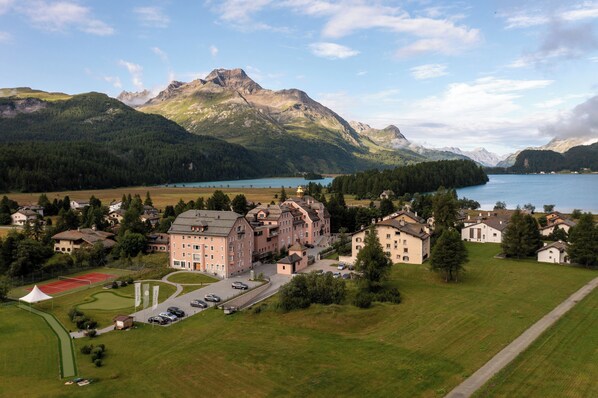 Paisagem montanhosa com um lago, quadra de tênis e um edifício grande com vários andares e uma torre central.