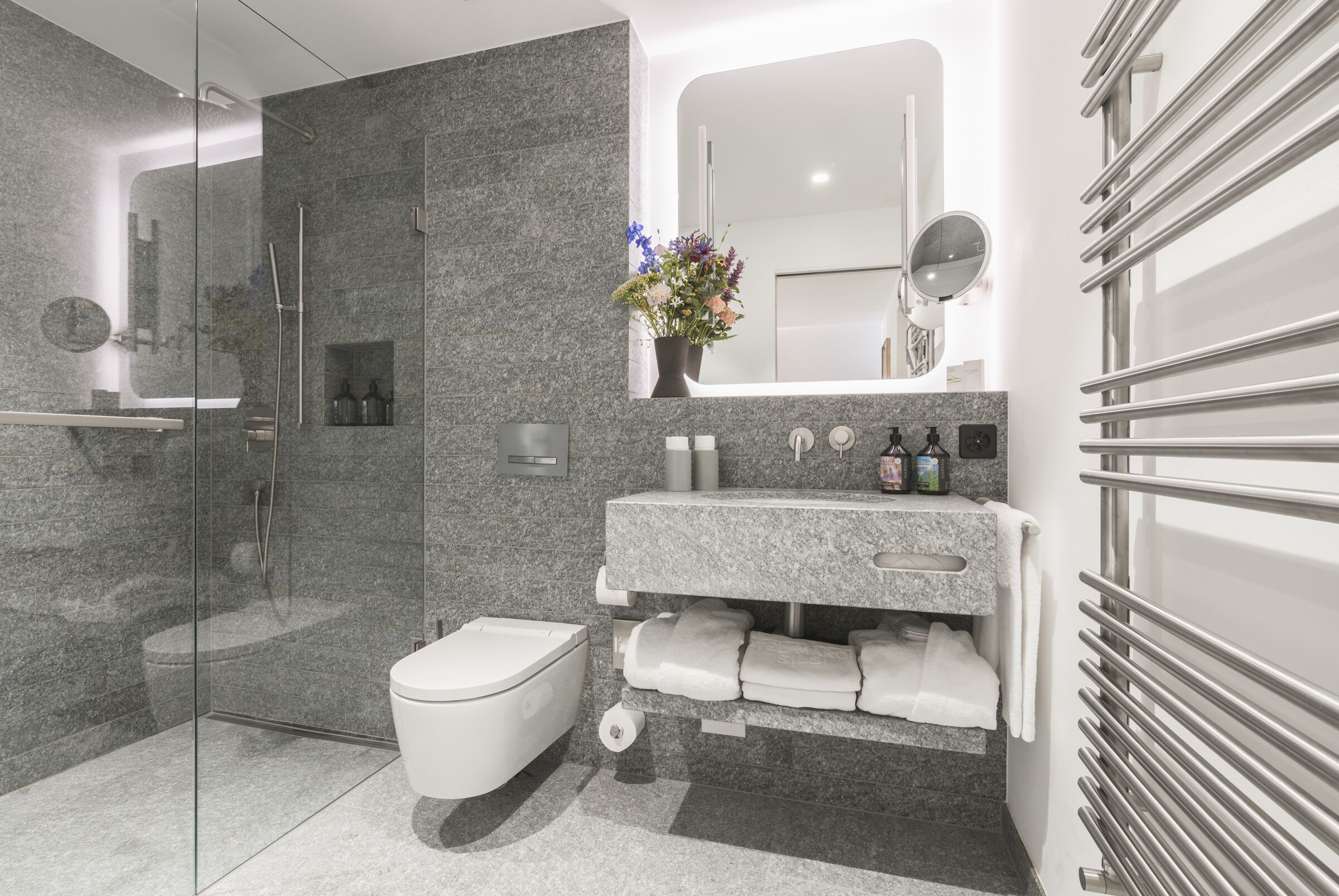 A modern bathroom with a glass-enclosed shower, a white toilet, a granite countertop vanity with a mirror, and a vase of flowers.