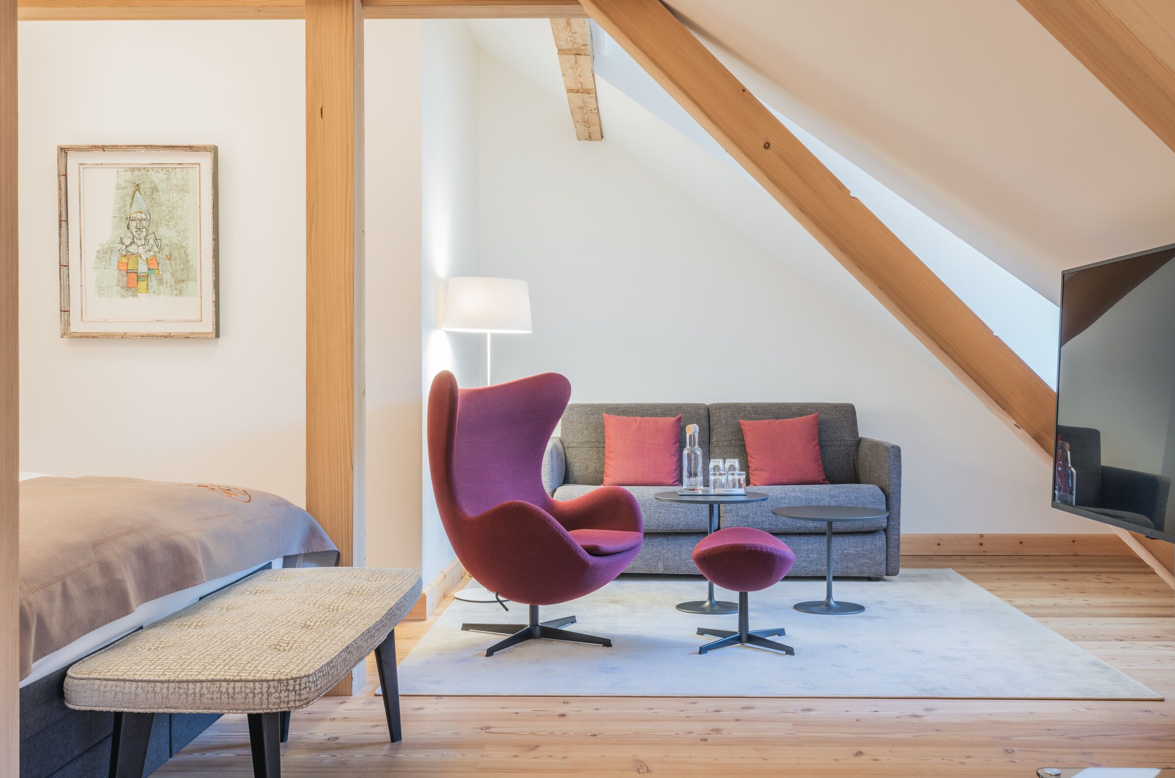 Un dormitorio tipo loft moderno con techo inclinado, una cama, un banco, un sillón morado, una mesita, un sofá y un televisor.