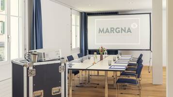 A conference room with a long table, chairs, a projector screen, and a presentation about Margna Parkhotel Islington.