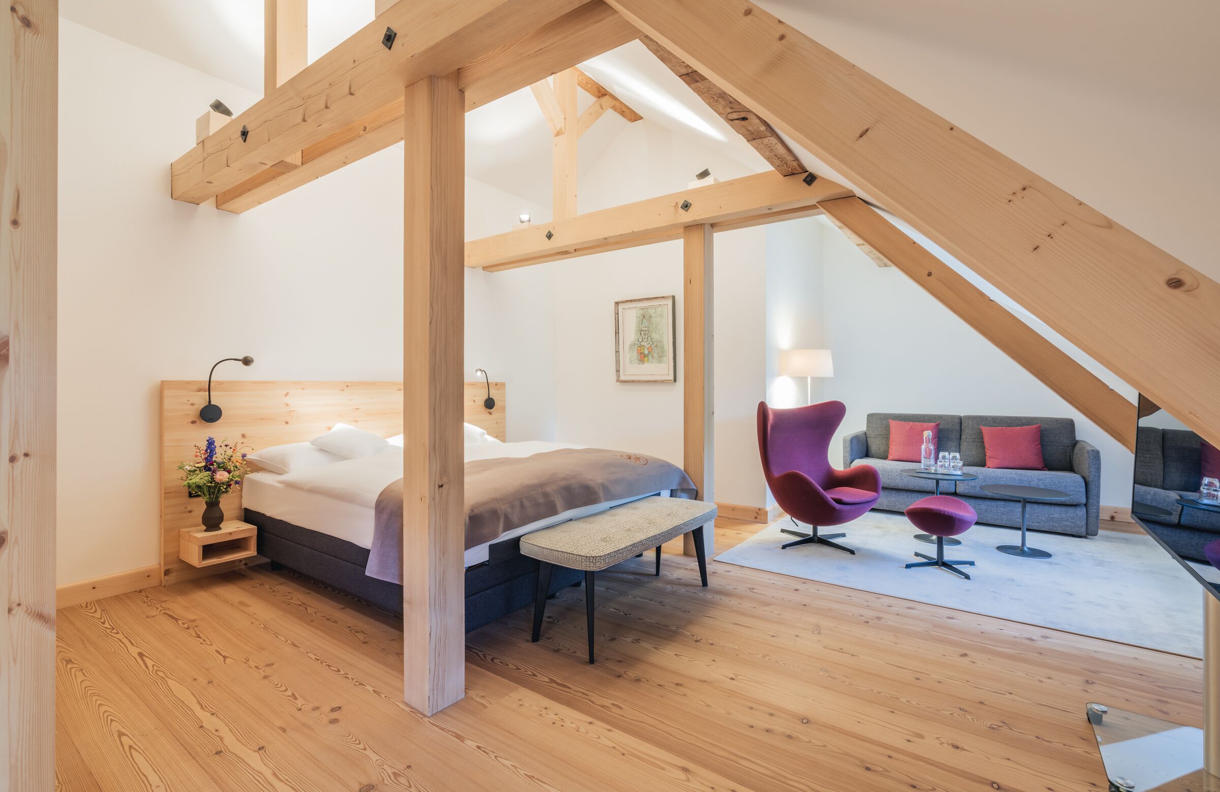 Ein Schlafzimmer mit Bett, Holzbalken, einer Bank, einem Sessel und einer Couch in einem Loft-Zimmer.