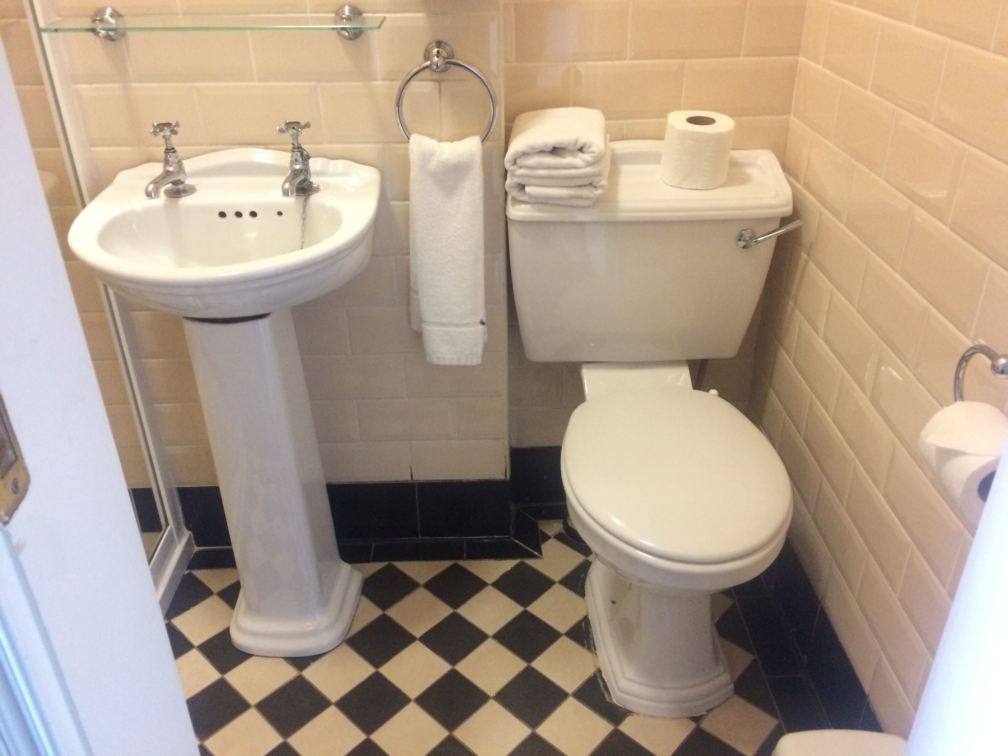 Un baño con lavamanos pedestal, inodoro y repisa de vidrio sobre el lavamanos.