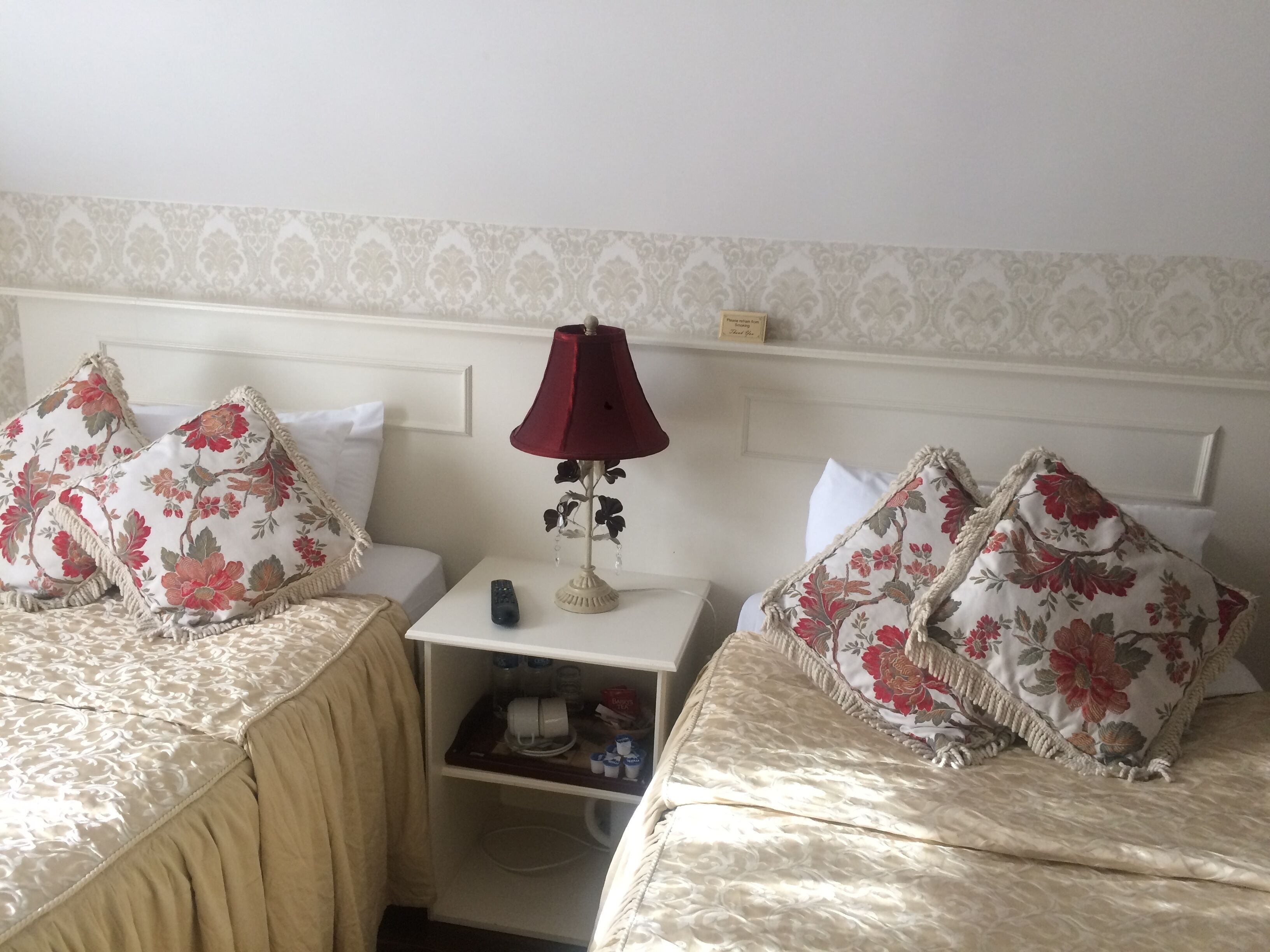 A hotel room with two beds, each with floral-patterned pillows and a red lamp on a bedside table.