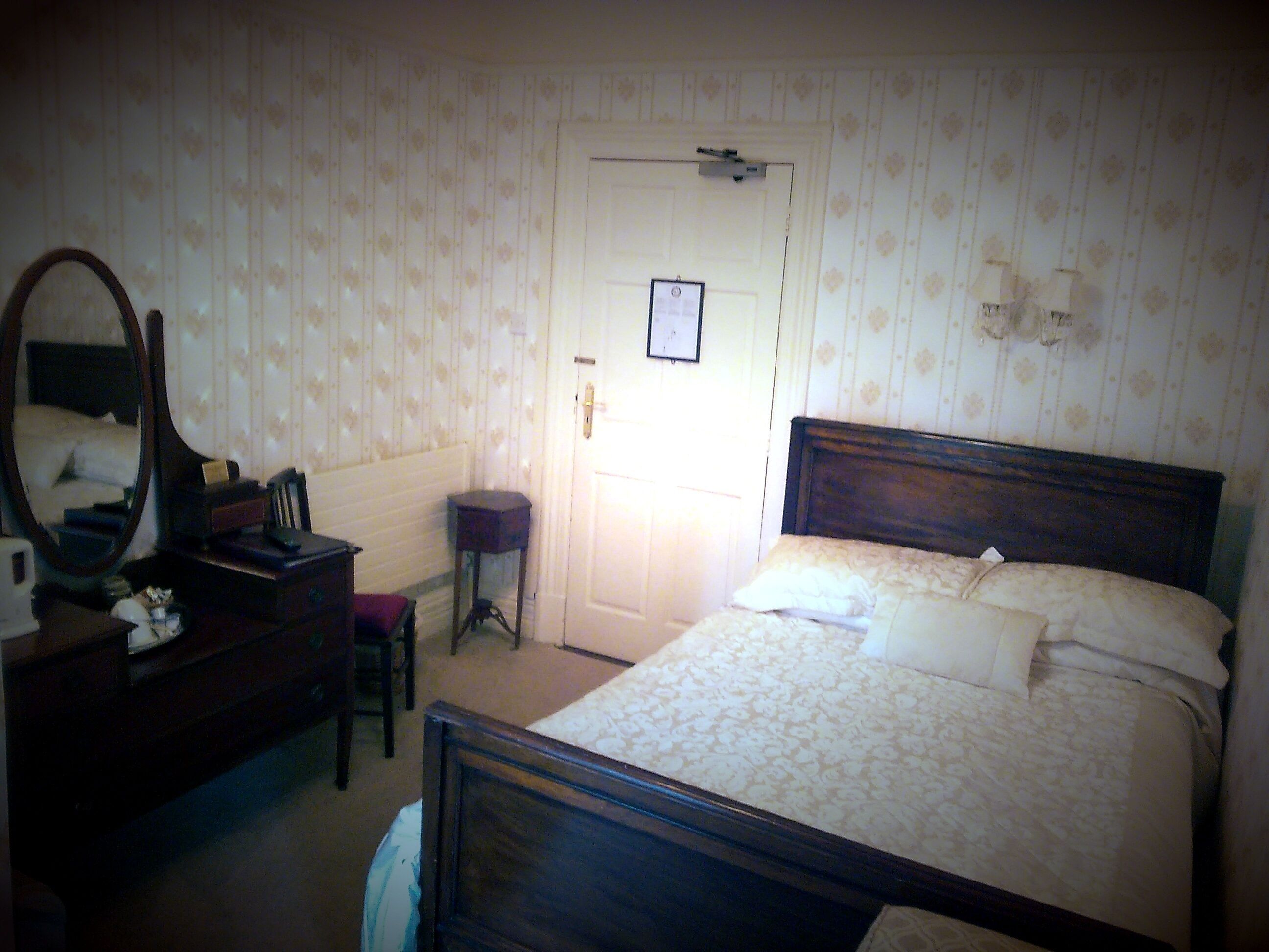 A bedroom with a wooden bed, a bedside table, a mirror, and a door.