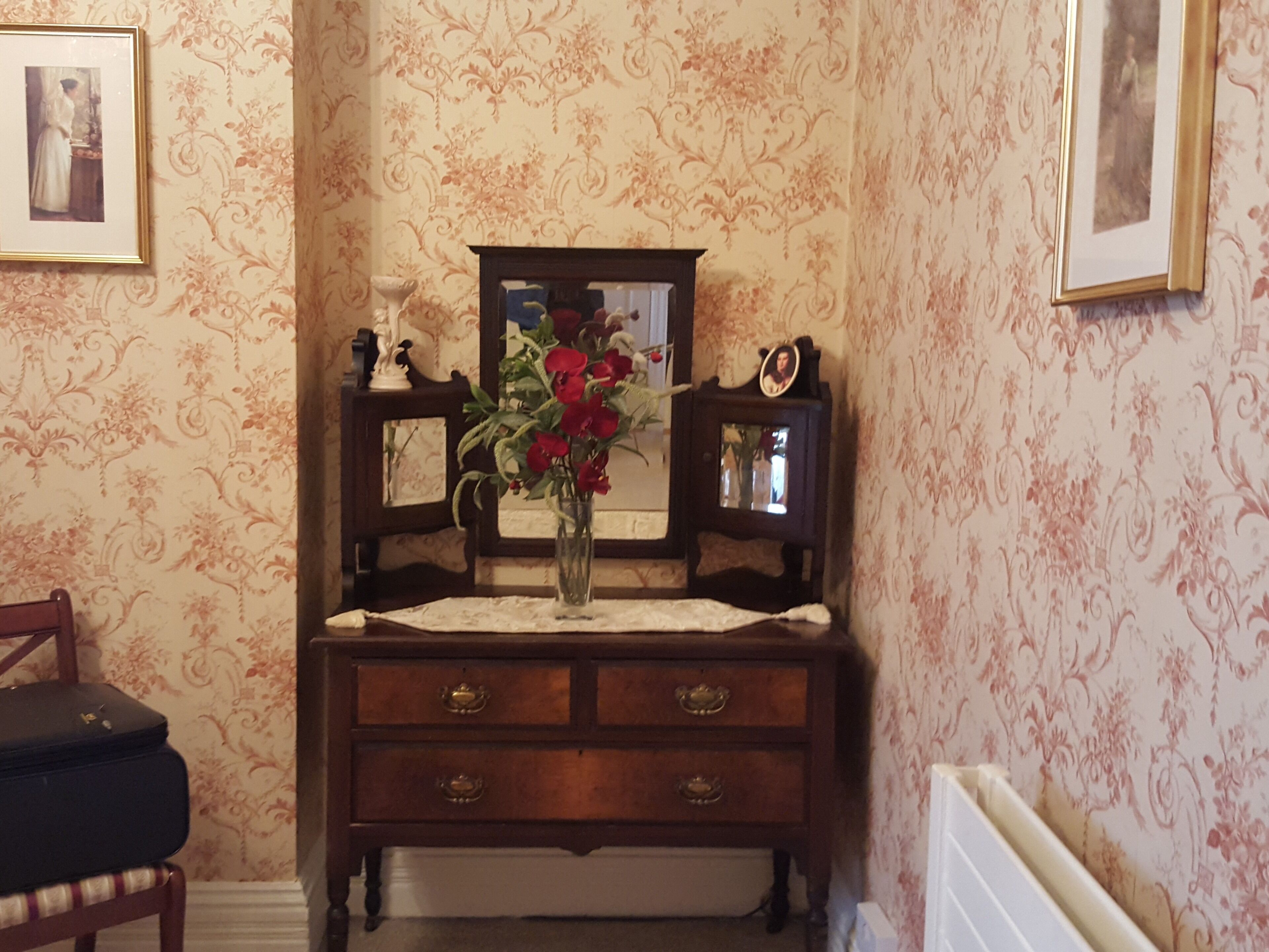 A room with a wooden dresser, a mirror, and a vase with flowers. There is a framed picture on the wall and a small table with a vase of red flowers.