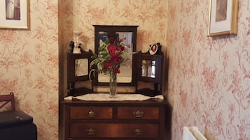 A room with a wooden dresser, a mirror, and a vase with flowers. There is a framed picture on the wall and a small table with a vase of red flowers.