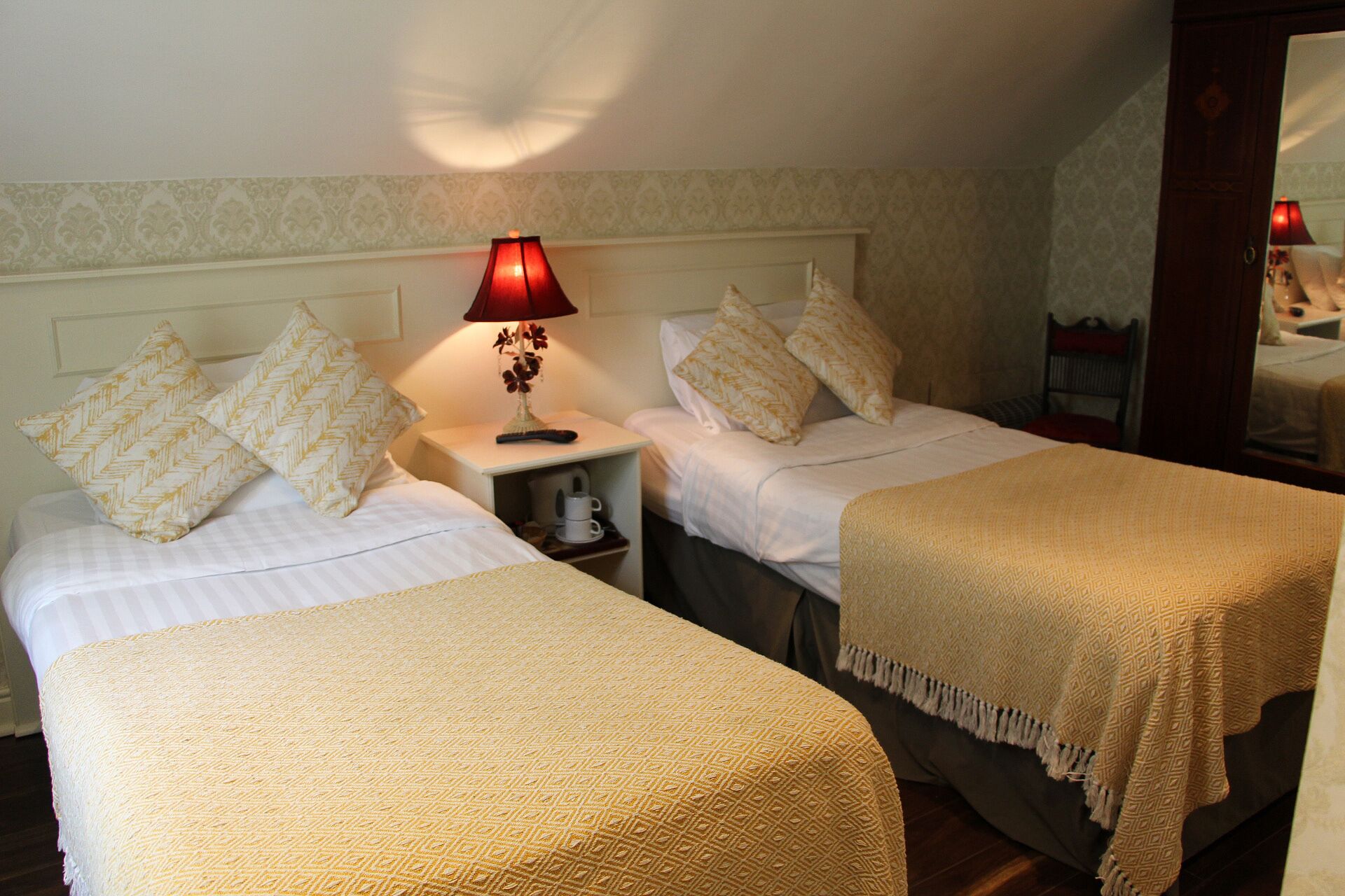 A hotel room with two single beds, each with a patterned pillow and a beige throw blanket. A red table lamp with a floral design is on the nightstand between the beds.