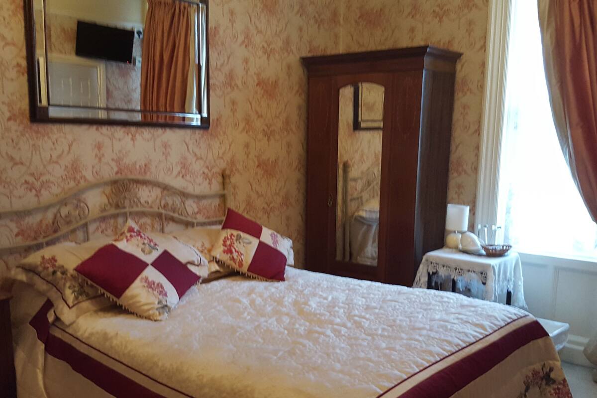 A bedroom with a bed, a wooden wardrobe, a small table with a lamp, and a mirror on the wall.