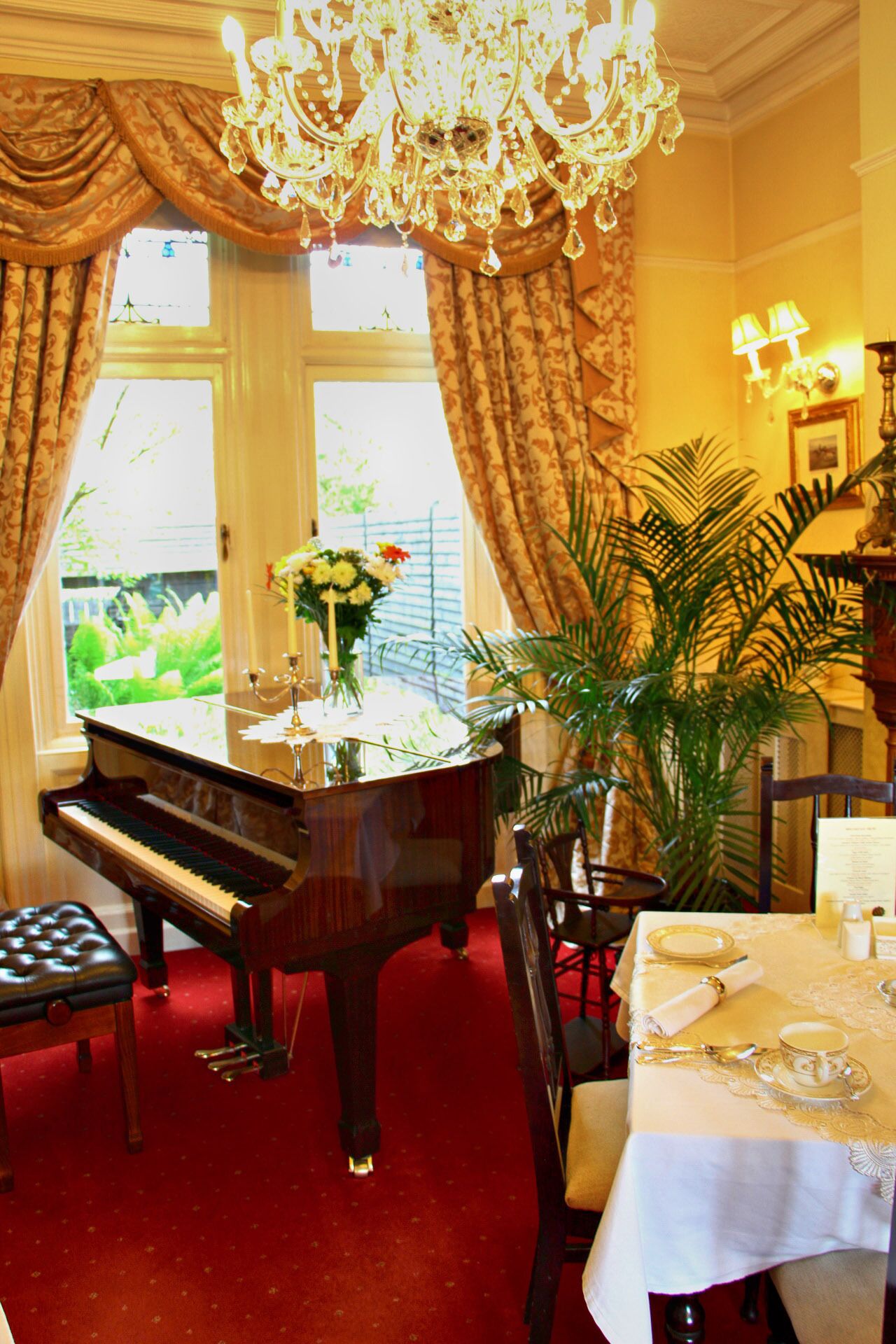 A grand piano in a room with a red carpet, a chandelier, and a dining table set with goldware.