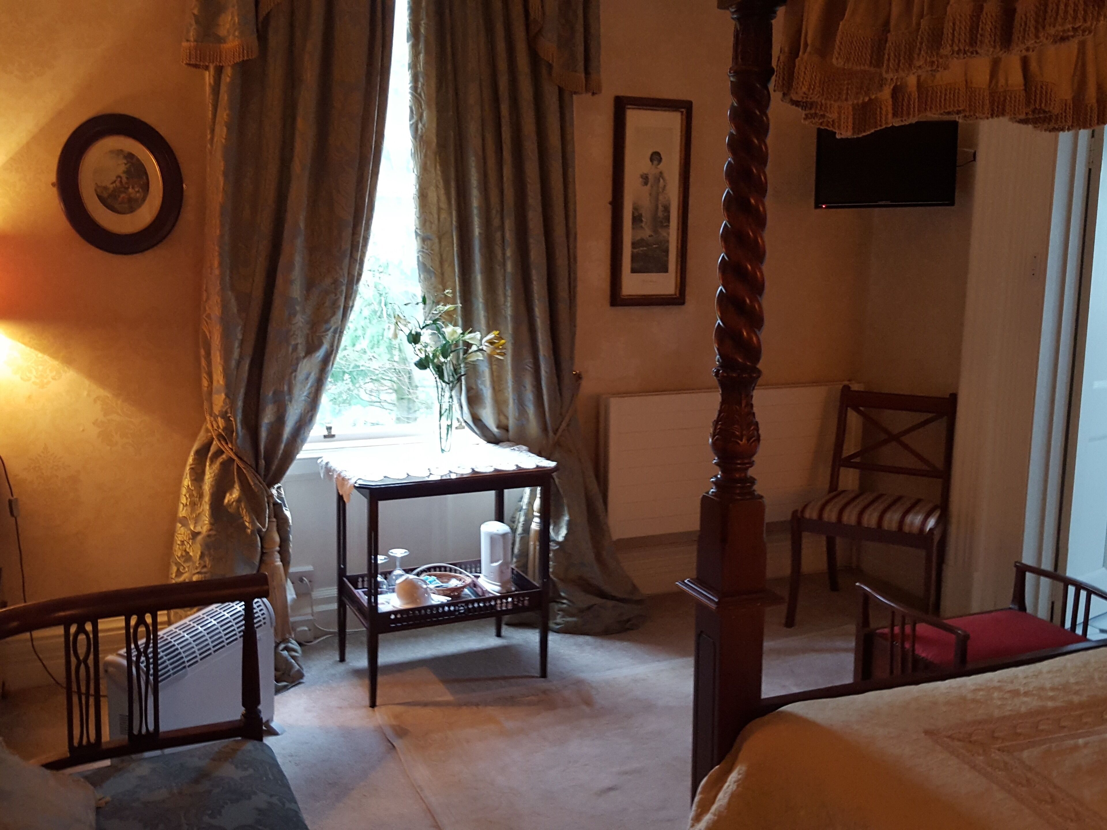 Une chambre à coucher avec un lit à baldaquin, une petite table sur laquelle est posé un vase, une chaise et un radiateur.