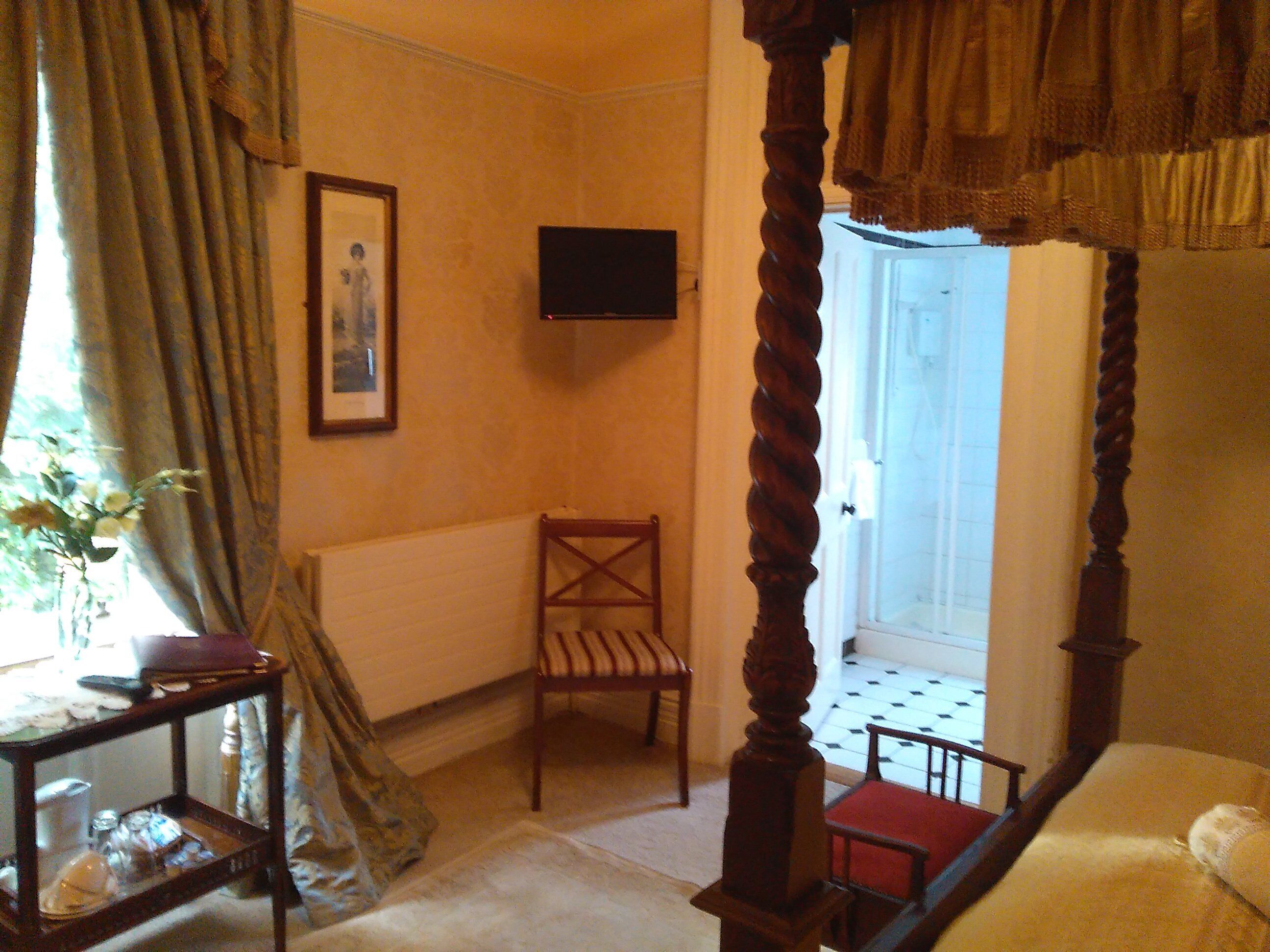 A bedroom with a four-poster bed, a television on the wall, a chair, and a small table with a vase of flowers.
