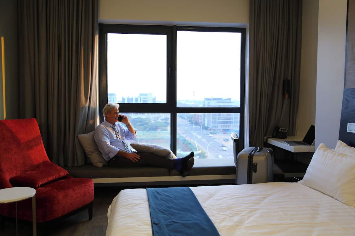 A hotel room with a large window, a red armchair, a bed with white and blue bedding, and a desk with a laptop.