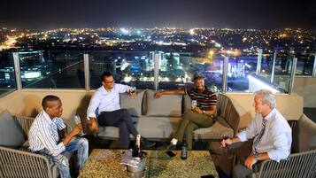 Un groupe de personnes est assis sur une terrasse en toit, d’où l’on a une vue sur la ville. Sur la table, on peut voir une bouteille de champagne et une bouteille de vin.