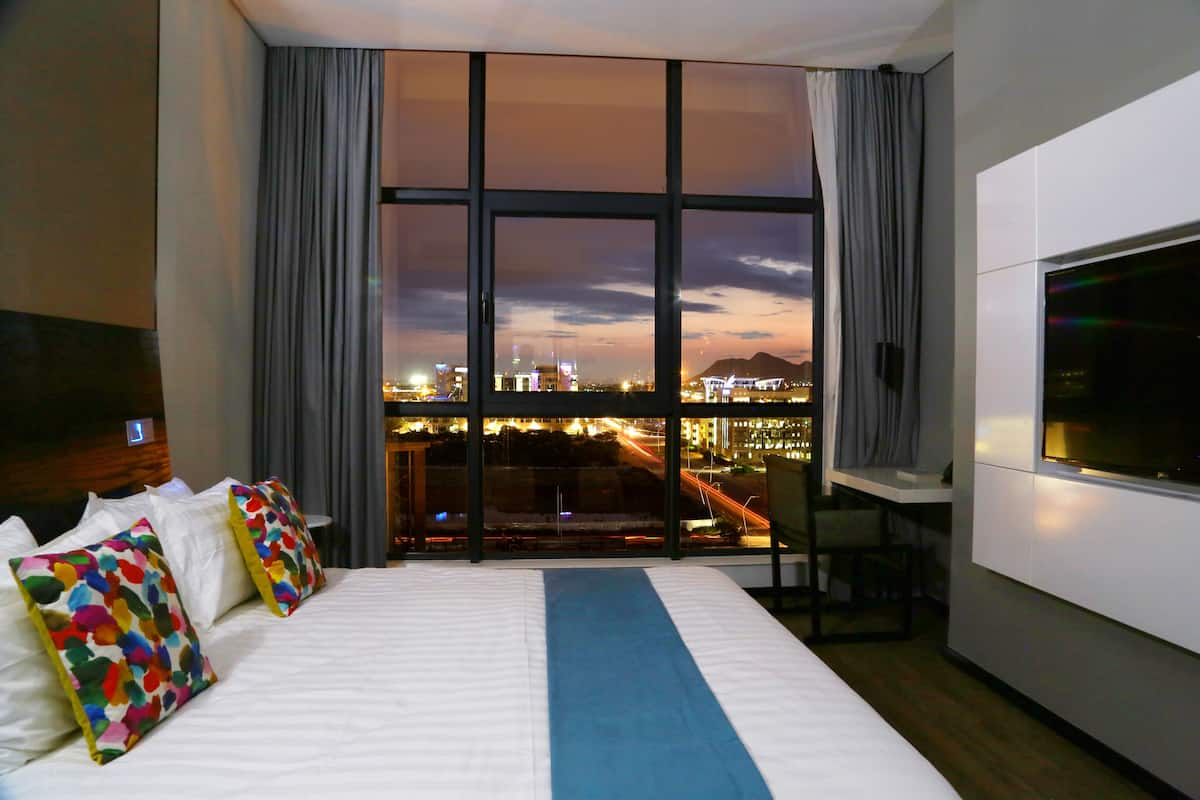 A hotel room with a large bed, a TV, a desk, and a view of a cityscape at dusk.