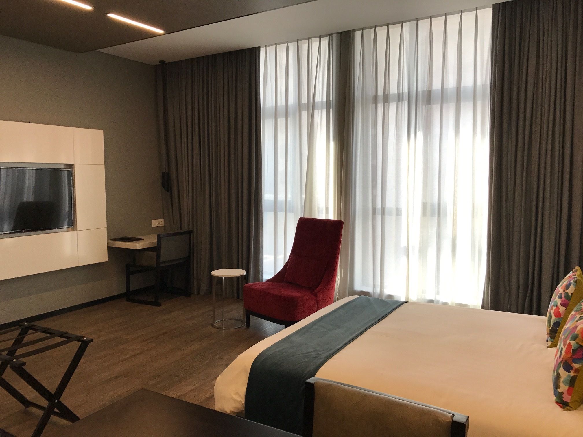 A hotel room with a bed, a TV mounted on the wall, a desk with a chair, and a red armchair.