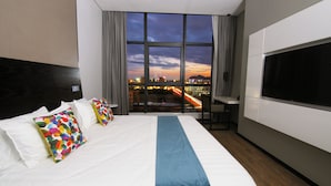 A hotel room with a large bed, a TV, and a view of a cityscape at dusk.