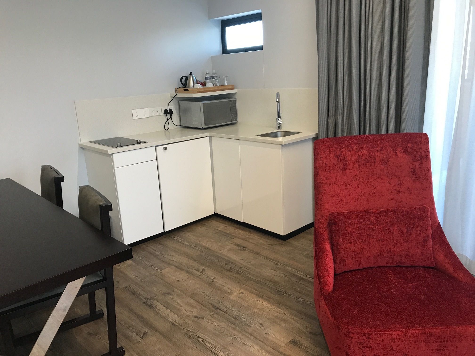 A compact kitchen with white cabinets, a microwave, and a sink. There's a red chair and a dark wooden table.