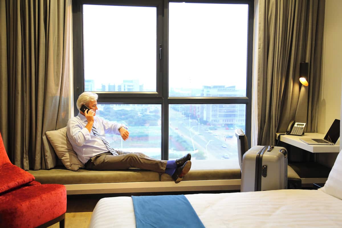 A hotel room with a large window, a bed, a red chair, a desk with a laptop, and a suitcase.