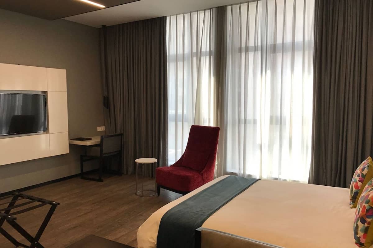 A hotel room with a bed, a desk, a chair, a TV mounted on the wall, and large windows with curtains.