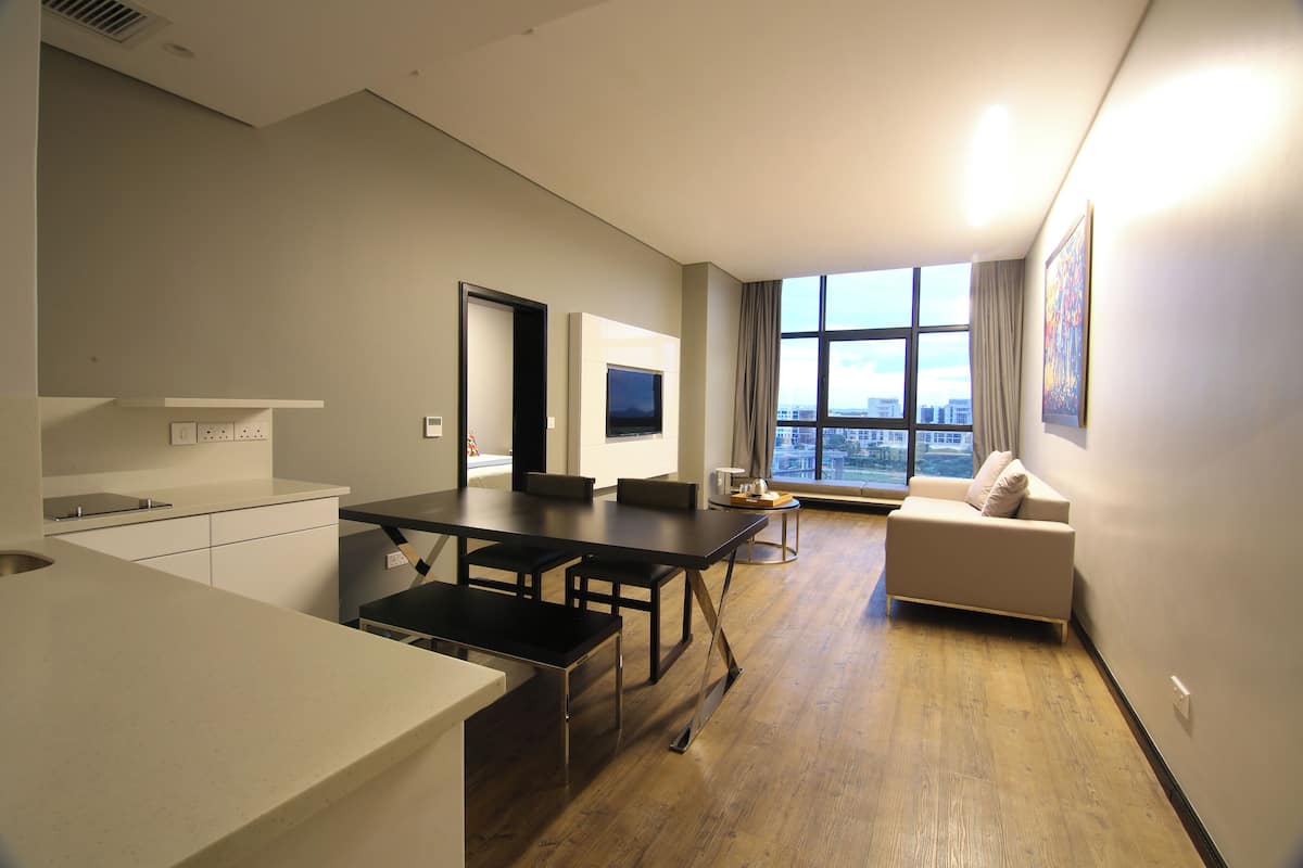 A modern apartment with a kitchen, dining area, and living room. It features a large window with a city view, a flat-screen TV, and a sofa.