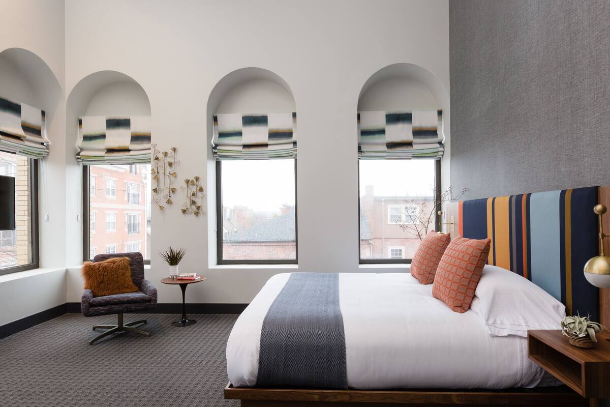 Une chambre d’hôtel moderne, dotée d’un grand lit, d’une chaise, d’une petite table et de trois fenêtres en arc surmontées de rideaux à motifs.