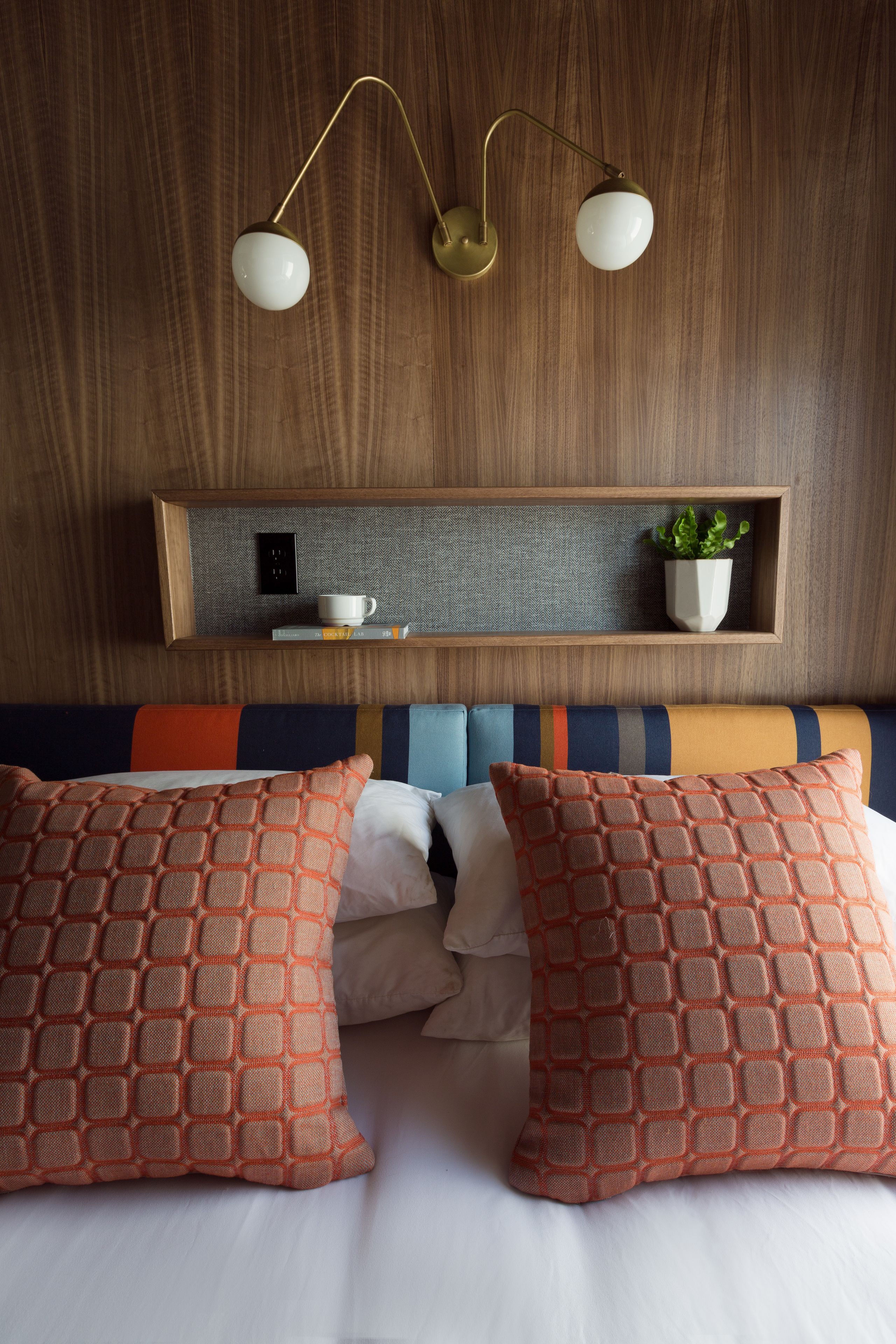 Habitación de hotel con cabecera de madera, una mesita de noche con una taza y una planta, y una cama con almohadas estampadas color naranja.