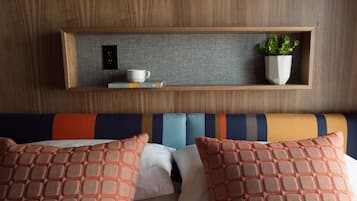 Habitación de hotel con cabecera de madera, una mesita de noche con una taza y una planta, y una cama con almohadas estampadas color naranja.