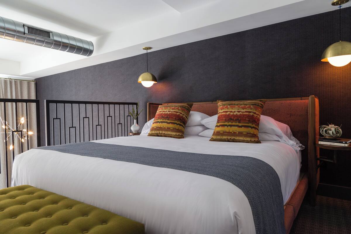 Un lit bien fait, avec une couette grise et blanche, deux coussins à motifs et un pouf vert. On y trouve une table de chevet avec une lampe et un petit objet décoratif. La pièce a un mur de couleur sombre et une bouche d’aération au plafond.